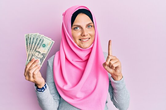 Young caucasian woman wearing islamic hijab holding 20 dollars banknotes smiling with an idea or question pointing finger with happy face, number one