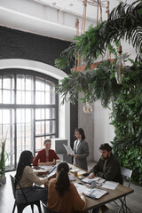 Vertical high angle portrait of diverse group of business people meeting at table in modern office interior decorated by plants, copy space