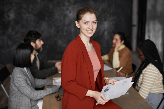 Waist Up Portrait Of Successful Young Businesswoman Wearing Red Jacket And Smiling At Camera With Diverse Group Of People Meeting In Background, Copy Space