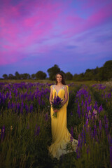 A beautiful girl in a long yellow dress against the background of a blooming purple lupine field and a bright sunset sky. 