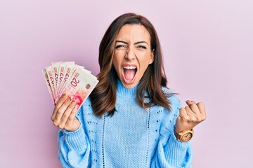 Young brunette woman holding 20 israel shekels banknotes angry and mad raising fist frustrated and furious while shouting with anger. rage and aggressive concept.