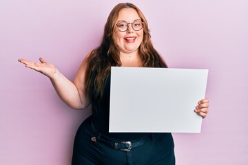 Naklejka premium Plus size caucasian young woman holding blank empty banner celebrating achievement with happy smile and winner expression with raised hand