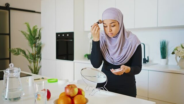 Young modern woman in hijab doing makeup in front of the mirror while sitting in the kitchen. Applies foundation with a brush