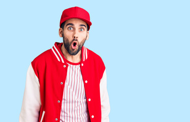 Young handsome man with beard wearing baseball jacket and cap scared and amazed with open mouth for surprise, disbelief face