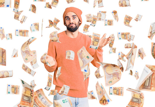 Young handsome man with beard wearing casual sweater and wool cap clueless and confused expression with arms and hands raised. doubt concept.