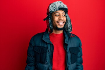 Young african american man with beard wearing winter hat and coat looking away to side with smile on face, natural expression. laughing confident.