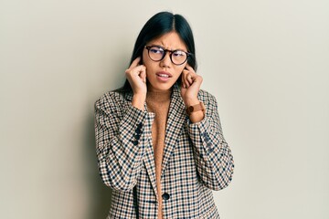 Young chinese woman wearing business style and glasses covering ears with fingers with annoyed expression for the noise of loud music. deaf concept.