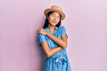 Young chinese woman wearing summer hat hugging oneself happy and positive, smiling confident. self love and self care