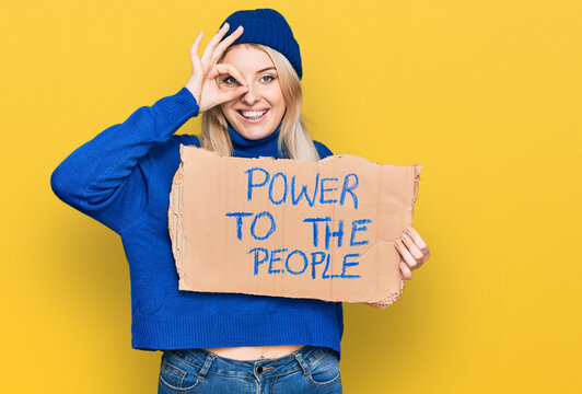 Young caucasian woman holding power to the people banner smiling happy doing ok sign with hand on eye looking through fingers