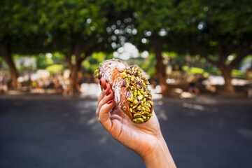 Female hand holding a pistachio cannoli in front of a park - obrazy, fototapety, plakaty