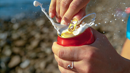 MACRO: Woman relaxing on the beach opens up a can of beer and gets sprayed.