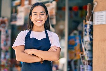 Young latin shopkeeper girl with arms crossed smiling happy at the press shop