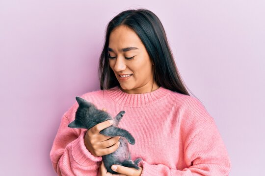 Young hispanic girl smiling happy holding cute cat over isolated pink background.