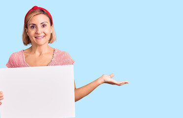 Young blonde woman holding blank empty banner celebrating victory with happy smile and winner expression with raised hands