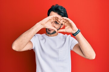 Young hispanic man wearing casual white tshirt doing heart shape with hand and fingers smiling looking through sign