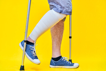 Legs of a man in right profile wearing shorts and on crutches, with a bandaged leg, on yellow background