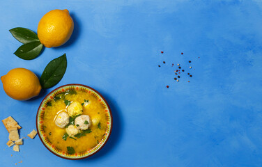 A bowl of avgolemono chicken soup, with meatballs. On a bright blue background with lemons, crackers and peppers. Top view with space