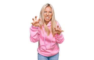 Young blonde woman wearing casual sweatshirt smiling funny doing claw gesture as cat, aggressive...
