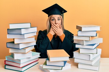 Young caucasian woman wearing graduation ceremony robe sitting on the table shocked covering mouth with hands for mistake. secret concept.