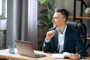 Handsome successful self-confident middle-aged caucasian business leader or manager, sitting in an office in a stylish formal suit, thinking about a project, looking away, taking notes, smiling