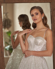 Fototapeta premium Portrait og young beautiful woman wearing white wedding bridel dress. Pretty slim bride with natural make-up and wavy hair posing against mirror