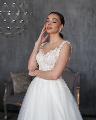 Fototapeta na wymiar Pretty young caucasian brunette bride with make-up and pony tail hairstyle in long wedding ball gown dress standing and posing in grey interior