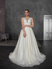 Fototapeta premium Pretty young caucasian brunette bride with make-up and pony tail hairstyle in long wedding ball gown dress standing and posing in grey interior