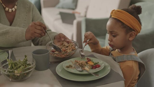 Medium shot of little African-American girl wearing casual clothes sitting at kitchen table and eating while unrecognizable African woman putting food on her plate