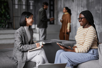 Portrait of two ethnic businesswomen discussing project while sitting in lounge area at modern office