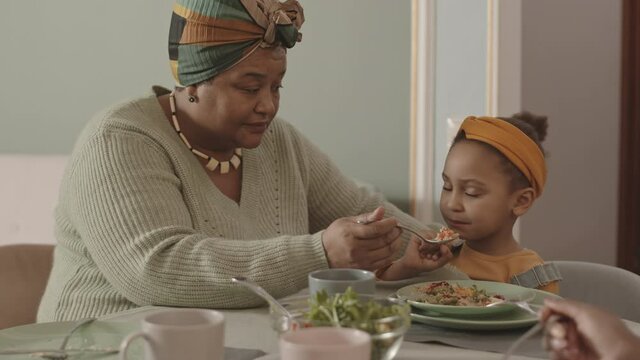 Medium shot of beautiful old African-American woman wearing casual clothes sitting at dining table and trying to feed her little granddaughter who refusing to eat - Powered by Adobe