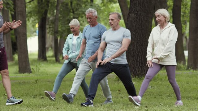 Lockdown of group of four aged sporty people doing forward lunges in park while handsome aged bearded trainer watching them
