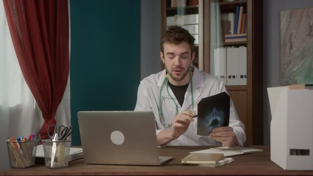 Confident young American doctor reading x-ray picture, concentrated facial expression, treating patient online using video chat internet connection on laptop computer