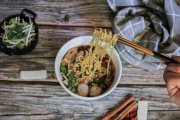Hand uses chopsticks to pickup tasty egg noodle with pork and vegetable in five spices black soup on wooden table top view- Asian food style.
