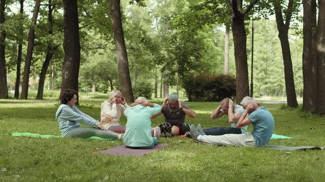 Wide shot of several aged sportsmen and sportswomen lying on mats on lawn in park and doing sit-ups together