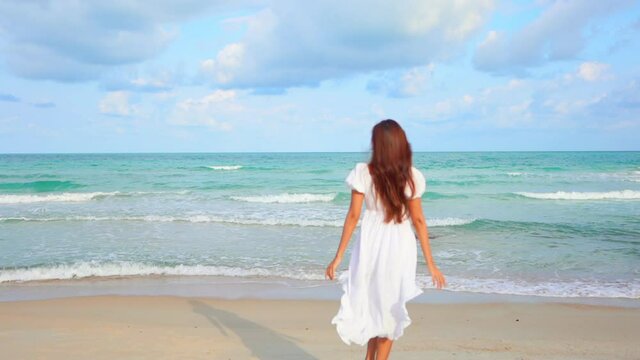 Back of attractive woman in fluttering summer dress walking on sand beach by sea looking at skyline and raising hands, slow motion