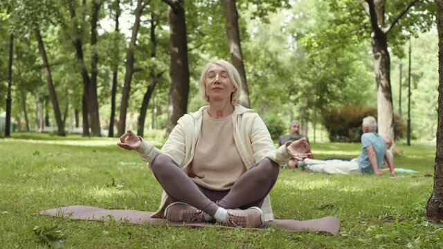 Wide shot of aged Caucasian woman with blond hair wearing sportswear sitting in lotus position on yoga mat in park, meditating then looking at camera and smiling