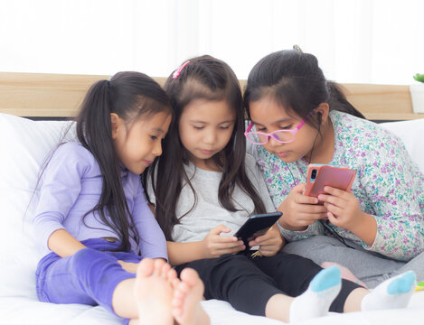 Happy Asian Little Kid And Friends Playing Smartphone On Sofa At Home, Children Using Phone Together On Couch, Girl Watching Smart Phone For Entertainment, Lifestyle And Communication Concept.