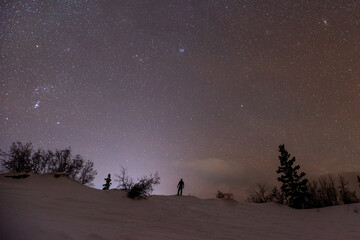 Naklejka na ściany i meble A starry, star filled night with a person standing on a snowy, snow covered ridge with bright sky above. Wilderness area with spruce trees and bushes. 