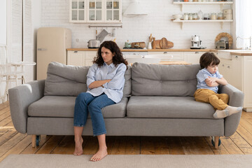 Fototapeta premium Annoyed mom and stubborn child sitting on couch at home, ignoring each other, posture of discontent. Sad mother disobedient kid son sitting apart on sofa with arms crossed, not talking after quarrel.