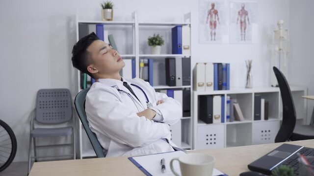 tired asian male medical worker is throwing back his head with closed eyes and folded arms while sitting resting in the office chair at a modern bright clinic.