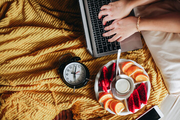 Hand typing on laptop keyboard with various fruit for breakfast and clock showing 7 o'clock on the bed