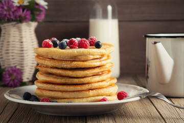 Stack of pancakes with fresh berries on a white plate, a bottle of milk and a vase of wildflowers on the table