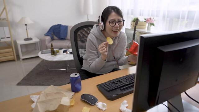 lazy girl indoorsy laughing and eat chips at home. asian chinese woman in headset and watching comedy show on internet with computer and enjoy snack. happy female homebody relax indoor on summer