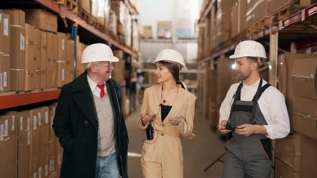 Young handsome man in work uniform and handsome woman in business suit talking to solid elderly man in warehouse with boxes