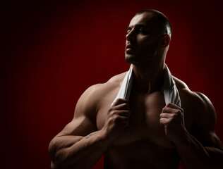 Strong muscular unshaved man, bodybuilder after workout stands in gym or shower with naked chest holding shirt or towel around his neck and looks aside at copy space on red background