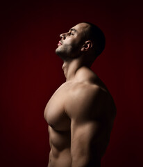Fototapeta premium Portrait of muscular man bodybuilder standing shirtless sideways toi camera with head up and looking at upper corner, taking rest after training over red background