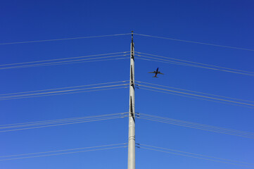 Commercial airplane flying near high-voltage power lines and utility pole against a clear blue sky, illustrating airspace activity intersecting with energy transmission infrastructure.