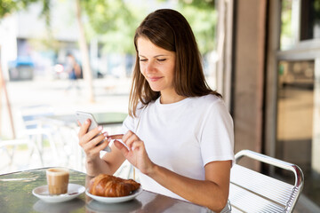 Cheerful young female using smartphone and enjoying coffee in city cafe