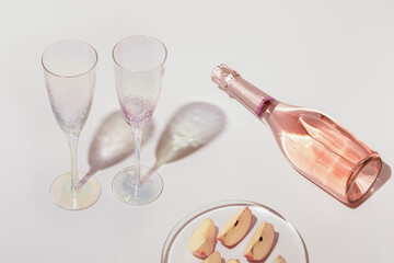 Сlosed bottle of rose champagne sparkling wine and glasses, pieces of apple fruit with sunlight on light table