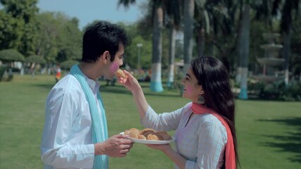 Beautiful woman feeding traditional Gujiya to an attractive male on Holi festival. Medium shot of youngsters celebrating the Indian festival of colors with sweet delicacies standing outdoors in the...